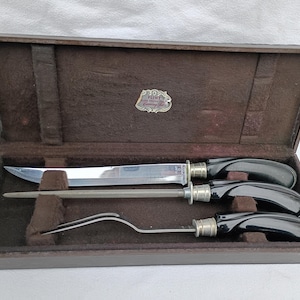 May include: A vintage carving set in a wooden case. The set includes a carving knife, a sharpening steel, and a carving fork. The handles are black with silver accents. The case has a brown exterior and a soft, brown interior.