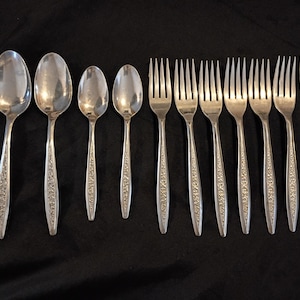 以下が含まれることがあります： 黒い背景に配置された、銀色のカトラリーのコレクション。スプーン、フォーク、ナイフが含まれています。これらの道具は、ハンドルに装飾的な花柄のデザインが施されています。スプーンはサイズが異なり、フォークには4つの歯があります。