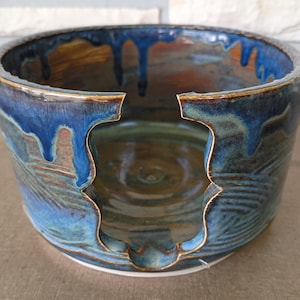 Może przedstawiać: Okrągła ceramiczna miska na włóczkę z głęboką niebieską glazurą, która ścieka po bokach na brązowo-beżową podstawę. Miska posiada szeroki, zakrzywiony otwór z przodu przeznaczony do prowadzenia włóczki oraz gładkie, błyszczące wykończenie.