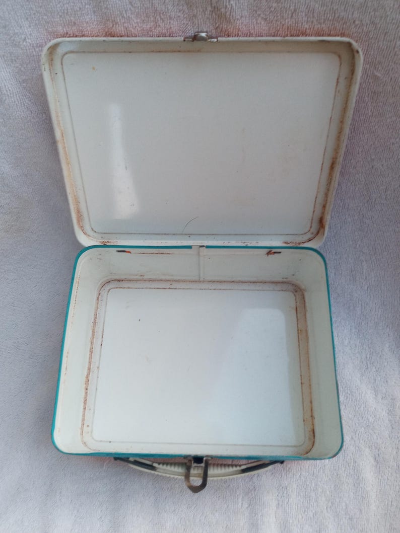 May include: An open, vintage metal lunchbox with a white interior and a turquoise blue exterior trim. The lunchbox has a hinged lid and a metal clasp closure. The edges show signs of rust, indicating its age.