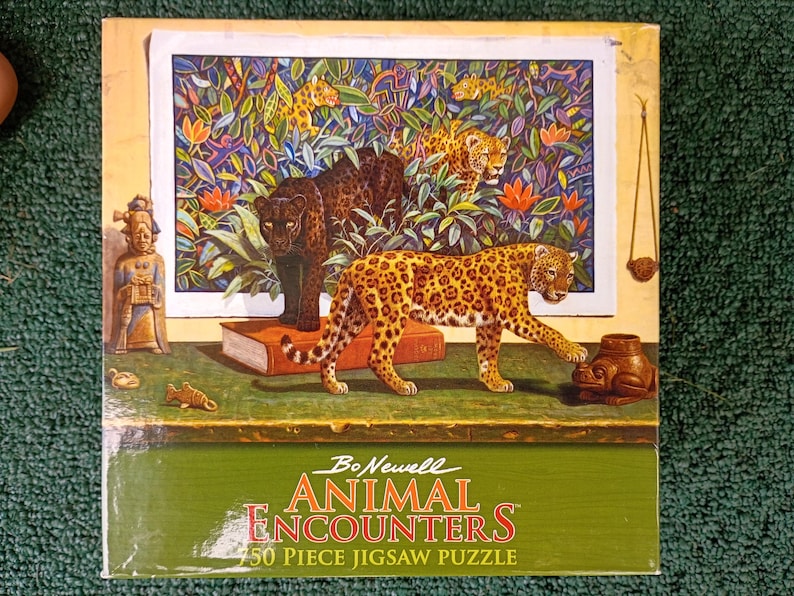 May include: A 750-piece jigsaw puzzle box titled Bo Newell Animal Encounters. The box features a painting of a black panther and a spotted leopard in a jungle setting, with a small figurine, a book, and a frog-shaped object on a table.