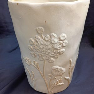 May include: A cream-colored ceramic vase with a textured surface and a slightly irregular rim. The vase features embossed floral designs, including a large flower and smaller botanical elements. The overall shape is cylindrical with a slight taper.