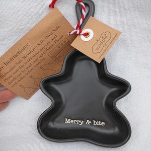 May include: Black ceramic gingerbread man-shaped dish with the text "Merry & bite". The dish has a loop for hanging and is attached to a tag with care instructions and a red and white string.