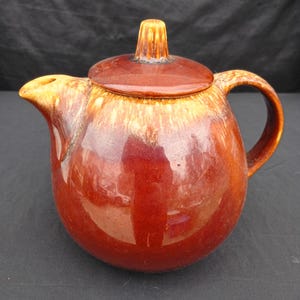May include: A brown ceramic teapot with a lid and handle. The teapot has a rounded shape and a glossy finish with a gradient of colours from light brown to dark brown. The lid has a small knob on top.