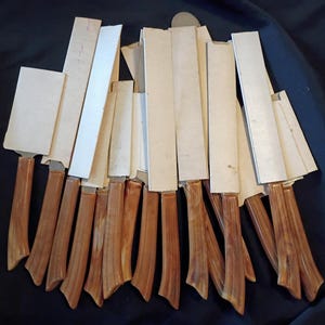 Puede incluir: Una colección de cuchillos con mangos de imitación madera en color marrón claro y fundas protectoras rectangulares blancas. Los cuchillos están dispuestos en grupo, con hojas metálicas dentro de fundas de cartón y mangos ergonómicos.
