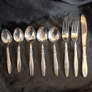 Può includere: Un set di posate in acciaio inossidabile, tra cui cucchiai, forchette e un coltello. I cucchiai hanno coppette ovali e manici decorativi. Le forchette hanno quattro rebbi e il coltello ha una lama arrotondata. Le posate sono disposte su uno sfondo scuro.