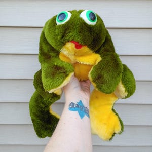 May include: A plush frog toy with large, round, green eyes and a red tongue. The frog is primarily green with yellow accents on its belly and legs. The toy is soft and fuzzy.