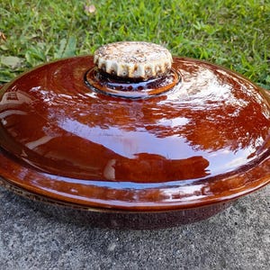 May include: A round, glossy, dark brown ceramic lid for a casserole dish. The lid features a smooth, reflective surface and a central, textured, off-white knob with brown speckles, designed for easy gripping and lifting during cooking tasks.