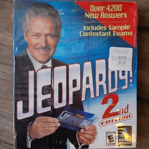 May include: A sealed box of the Jeopardy! 2nd Edition game. The box features a photo of the game show host, Alex Trebek, and the game's logo. The box also includes text that reads "Over 4,200 New Answers" and "Includes Sample Contestant Exams."