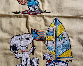 May include: Embroidered Snoopy designs on a light yellow fabric. One Snoopy is playing baseball, another is holding a flag, and a third is sailing a boat. The sailboat has a blue and yellow sail. The design includes the text "CUFS".