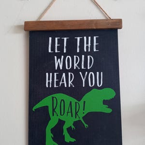 Pode incluir: Um quadro de parede preto com o texto "LET THE WORLD HEAR YOU ROAR!" em branco e uma ilustração de dinossauro verde. A placa é emoldurada com barras de madeira marrom e suspensa por uma corda natural.