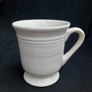 Puede incluir: Una taza de cerámica blanca con asa y base de pedestal. La taza tiene estrías horizontales alrededor del cuerpo y un asa curvada. La taza está sobre un fondo oscuro.
