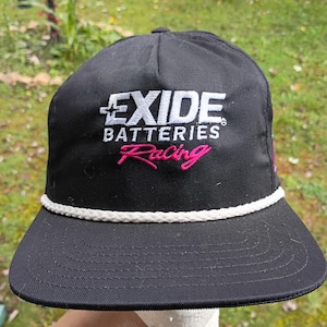 May include: A black baseball cap featuring the embroidered white text EXIDE BATTERIES with the word Racing in pink script below. The cap includes a white braided rope trim across the base of the crown where it meets the flat brim, with stitching.