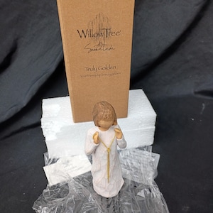 Puede incluir: Una figura de Willow Tree de una persona con un vestido blanco y un collar dorado. La figura está frente a una caja marrón con el texto "Willow Tree" y "Truly Golden". La caja está sobre una base de espuma blanca.