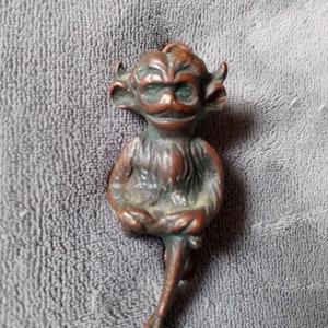 May include: A small, bronze figurine of a sitting, mischievous-looking creature with horns, a long tail, and a mischievous grin.