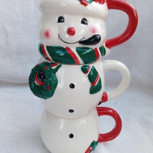 Peut inclure: Un ensemble de trois tasses en céramique blanche en forme de bonhomme de neige. Les tasses sont empilées les unes sur les autres pour former un bonhomme de neige. La tasse du haut a un chapeau rouge avec du houx vert, une écharpe rayée verte et rouge et un tuyau noir. La tasse du milieu a une couronne de Noël verte dans une main et une écharpe rayée verte et rouge. La tasse du bas a une poignée rouge et une branche de houx verte.