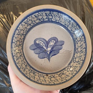 May include: A round ceramic plate with a blue floral design in the centre. The plate has a light beige background with a blue speckled border. The floral design is a stylized blue flower.