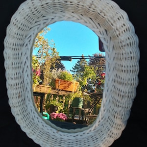 Könnte beinhalten: Weißer ovaler Spiegel aus Weidengeflecht mit geflochtenem Muster. Der Spiegel spiegelt einen Blick auf einen Garten mit grünen Bäumen und Pflanzen wider.