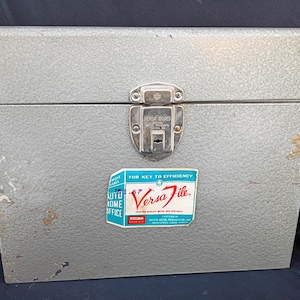 Puede incluir: Una caja Versa File de metal gris con cierre de pestillo. La caja tiene una superficie texturizada y una etiqueta que dice "Versa File" y "For Key to Efficiency". La etiqueta también dice "Auto Home Office".