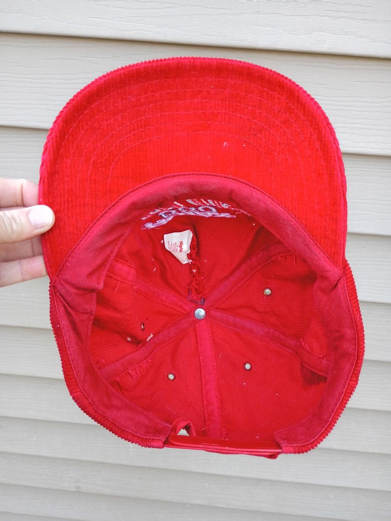 May include: A bright red corduroy baseball cap, viewed from the inside. The cap features a structured crown and a curved brim. The interior shows stitching and a small tag. The adjustable strap is visible at the back.