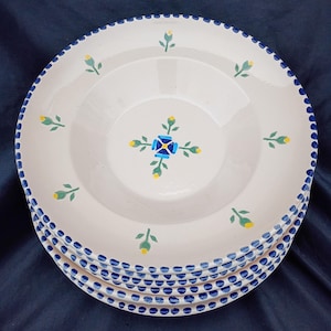 May include: A stack of white ceramic plates with a blue dotted border. Each plate features a central floral design in blue and yellow, with additional floral accents. The plates are stacked on a dark blue surface.