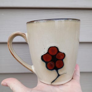 May include: Cream ceramic mug with a brown rim and handle. The mug features a stylized red and black floral design. The mug is held in a hand, showcasing its size and shape. A unique and artistic piece for enjoying beverages.