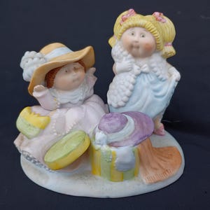 May include: A ceramic figurine featuring two Cabbage Patch Kids dolls. One doll is seated, wearing a pink dress and a yellow hat. The other doll is standing, wearing a blue dress and a white shawl. A purple hat is on the ground.
