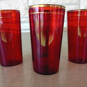 May include: Three cylindrical drinking glasses made of translucent red glass. Each glass features a smooth, straight-sided design with a thin, horizontal gold-coloured band encircling the upper rim area, providing a subtle metallic accent.