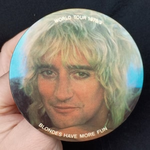 May include: Circular button with a portrait of a man with blonde hair. The button has the text "WORLD TOUR 1978/9" and "BLONDES HAVE MORE FUN". The background is a gradient of blue and brown.