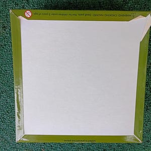 May include: A square cardboard box with a plain white center and a wide olive green border. The top edge features a small circular warning icon and the text: WARNING: CHOKING HAZARD - Small parts. Not for children under 3 years of age.