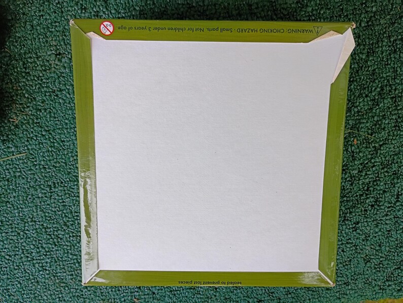 May include: A square cardboard box with a plain white center and a wide olive green border. The top edge features a small circular warning icon and the text: WARNING: CHOKING HAZARD - Small parts. Not for children under 3 years of age.