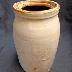 May include: A beige, antique-style ceramic jar with a wide opening and a slightly flared rim. The jar has a cylindrical shape with a rounded base and a textured surface. The rim is a darker shade of brown. The jar appears to be aged.