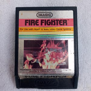 May include: A black Atari video game cartridge with a silver label. The label features the text "IMAGIC FIRE FIGHTER™" and "For Use with Atari® & Sears Video Game Systems". The image on the label shows a red fire truck driving through a fiery scene.