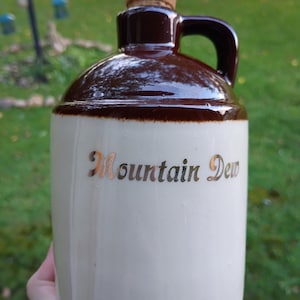 May include: A ceramic jug with a cream-colored body and a dark brown glazed top section. The jug features a single handle, a cork stopper, and the text Mountain Dew printed in gold lettering on the front of the cream-colored surface.