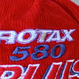 May include: Close-up of a red corduroy hat with embroidered text. The text includes "ROTAX" in white with a gold outline, "580" in blue, and partial text in red and white. The hat has a textured surface.