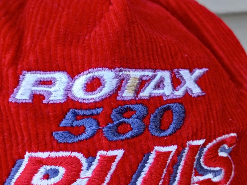 May include: Close-up of a red corduroy hat with embroidered text. The text includes "ROTAX" in white with a gold outline, "580" in blue, and partial text in red and white. The hat has a textured surface.
