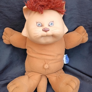 Peut inclure: Un chat en peluche vintage avec un corps, des bras et des jambes de couleur beige. Le chat a un visage beige clair avec des yeux bleus et un petit nez rose. Il a des cheveux roux et hirsutes et porte une tenue marron. Le jouet est en position assise, les bras tendus.