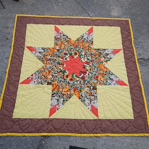 May include: A handmade quilt with a star design. The star's centre is a floral pattern with orange, yellow, and green hues. The star points are red and yellow, set against a yellow background, with a brown border.