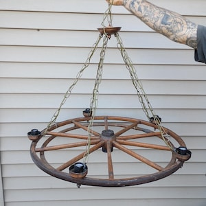 Puede incluir: Una lámpara de araña rústica con forma de rueda de carro con seis portalámparas. La rueda de madera está suspendida por cadenas de un dosel central. Los portalámparas son negros y cobrizos. El diseño es vintage y decorativo.