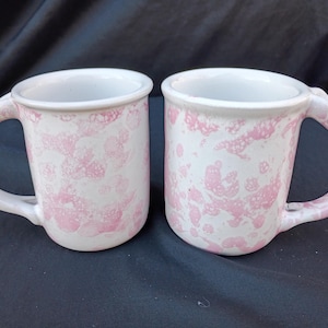 Può includere: Due tazze in ceramica bianca con un motivo marmorizzato rosa. Ogni tazza ha un manico arrotondato e un interno bianco. Le tazze sono esposte su una superficie blu scuro.