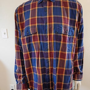 Puede incluir: Una camisa de cuadros de manga larga en azul, burdeos y amarillo. La camisa tiene dos bolsillos en el pecho y una botonadura. La tela parece ser de algodón o mezcla de peso medio. La camisa se exhibe en un maniquí.