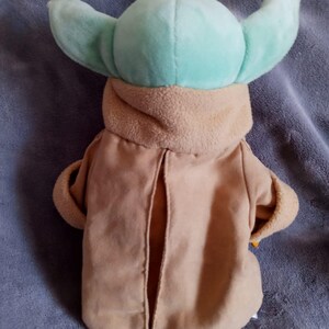 May include: A plush toy of a character with large, light green ears and a tan robe. The toy has a light green head and a tan-coloured robe with a slit down the front. The toy is designed to resemble a popular character from a science fiction franchise.