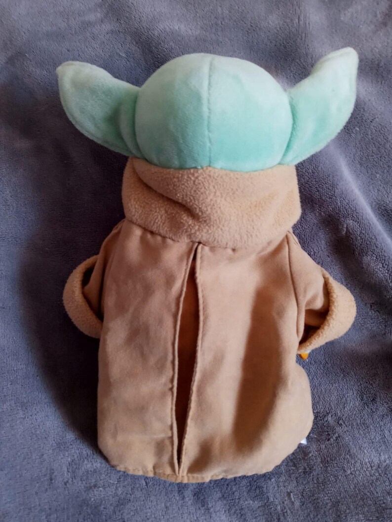 May include: A plush toy of a character with large, light green ears and a tan robe. The toy has a light green head and a tan-coloured robe with a slit down the front. The toy is designed to resemble a popular character from a science fiction franchise.