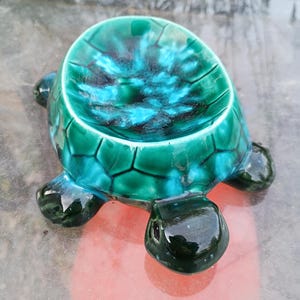 Puede incluir: Un plato de cerámica con forma de tortuga y acabado brillante. El caparazón de la tortuga es de un verde vibrante con detalles azules y negros, mientras que la cabeza y las patas son de un verde más oscuro. El plato probablemente se utiliza con fines decorativos.