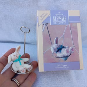 May include: A Hallmark Keepsake Easter ornament featuring a white bunny on a swing with a teal bow. The ornament is held by a silver metal stand. The box reads "Li'l Dipper" and "New Easter!"