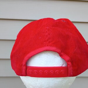 May include: A red corduroy baseball cap with a matching adjustable strap. The cap is displayed on a white foam head. The hat has a classic six-panel design.