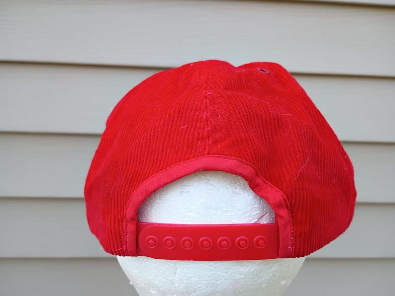 May include: A red corduroy baseball cap with a matching adjustable strap. The cap is displayed on a white foam head. The hat has a classic six-panel design.