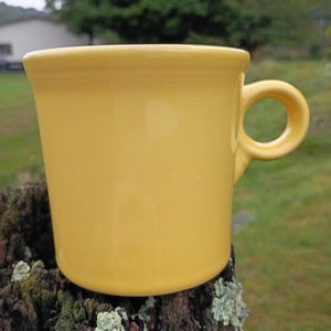 May include: A solid yellow ceramic mug with a smooth, glossy finish and a circular handle. The mug features a slightly flared rim and a cylindrical body, showcasing a vibrant, uniform yellow colour throughout the entire exterior surface.