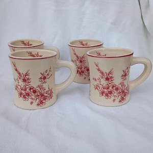 Może przedstawiać: Zestaw czterech kremowych ceramicznych kubków z czerwonymi kwiatowymi wzorami i czerwonym obramowaniem. Każdy kubek ma uchwyt i cylindryczny kształt. Kwiatowy wzór pokrywa korpus kubków.