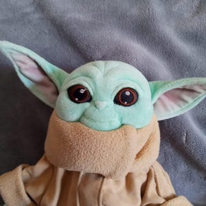 May include: A plush toy of a character with large ears, big brown eyes, and a light green face. The character is wearing a tan coat and a tan scarf. The toy is on a grey surface.